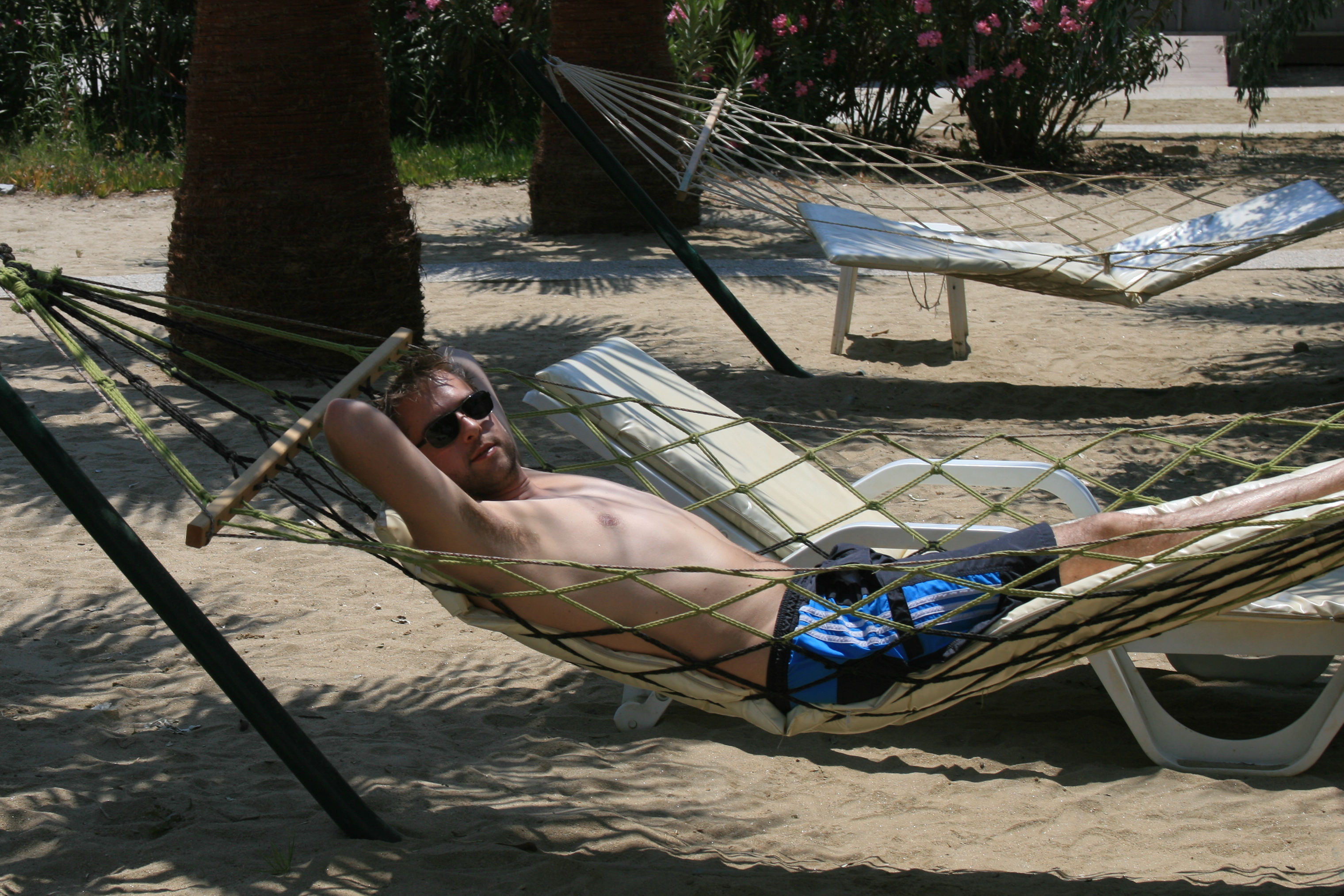 A man wearing sunglasses and swim shorts relaxes on a hammock on a sandy beach.