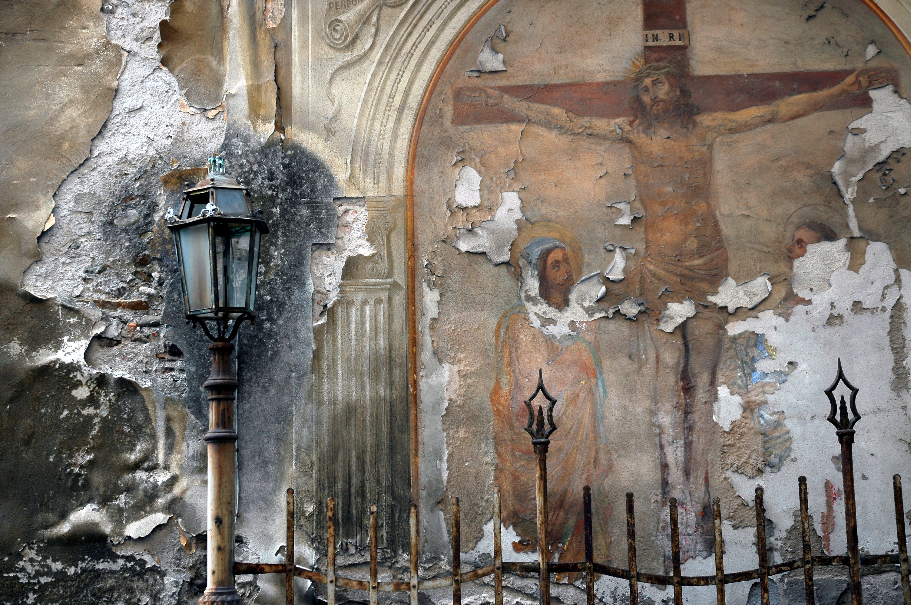A faded fresco on a damaged wall depicts Jesus on the cross with two figures below, partially peeling away.
