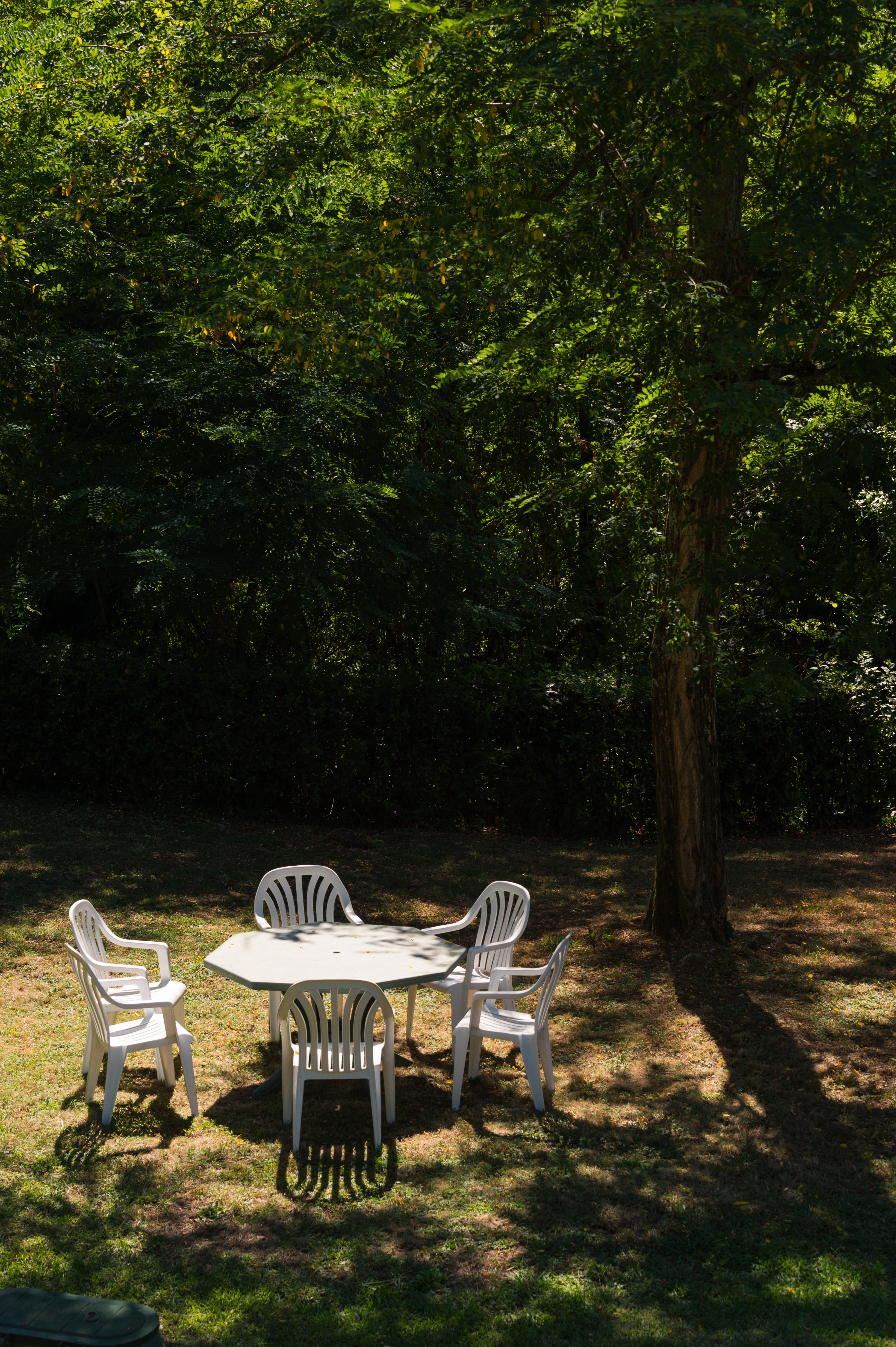 A white plastic table with four matching chairs sits on grass under the shade of a tree.