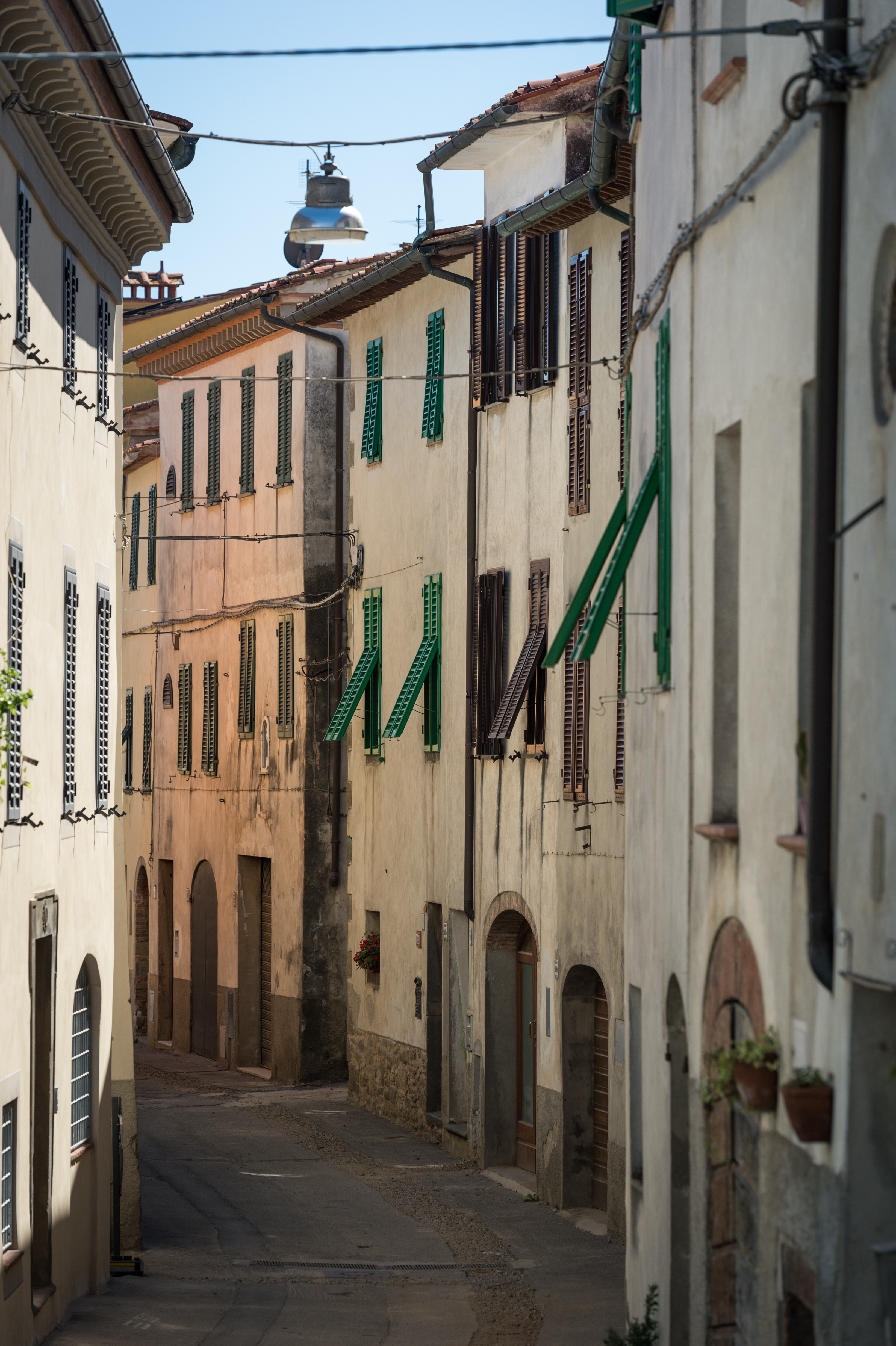 Narrow Italian street lined with old buildings, green shutters, and arched doorways, with a hanging street lamp above.