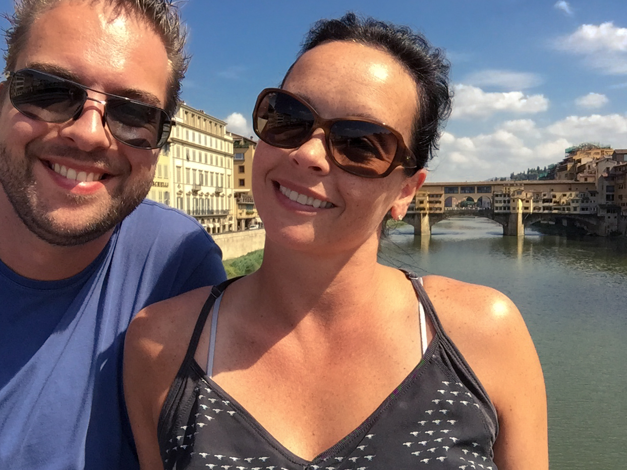 Two people wearing sunglasses take a selfie with the Ponte Vecchio bridge in the background on a sunny day.