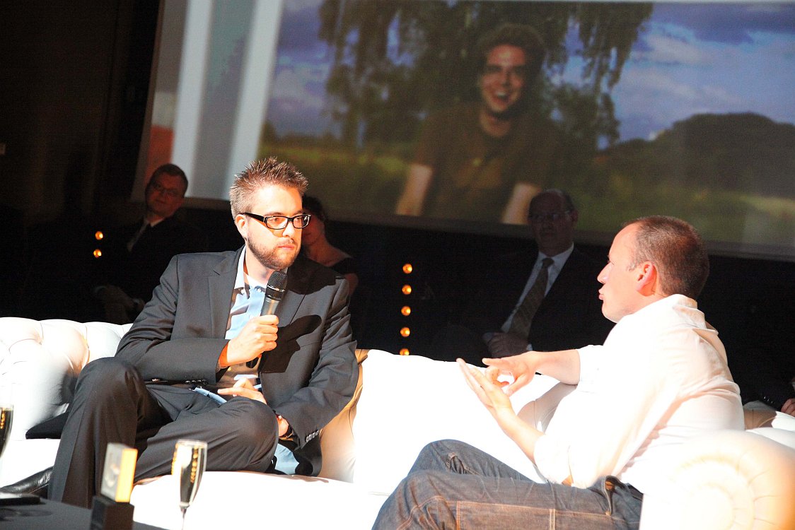 A man in a suit holds a microphone while speaking with another man during an alumni event.