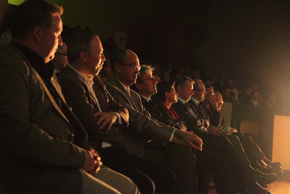 A group of formally dressed people sits in an audience, attentively watching an illuminated stage or presentation.