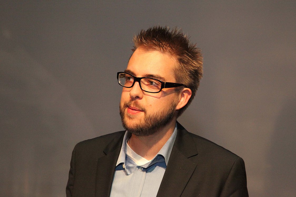 A man in a suit and glasses looks to the side, possibly during an event or presentation.