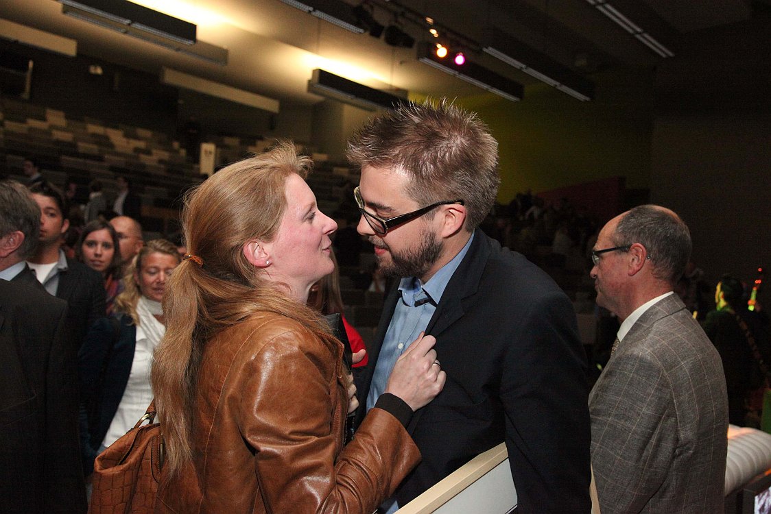 A man holding a certificate is congratulated by a woman in a crowded auditorium.