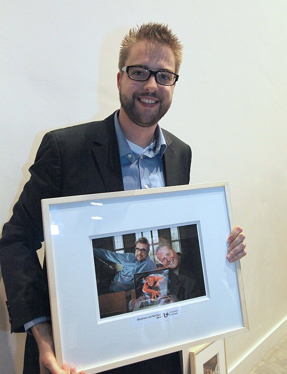 A man in a suit smiles while holding a framed award photo labeled "Alumnus of the Year.