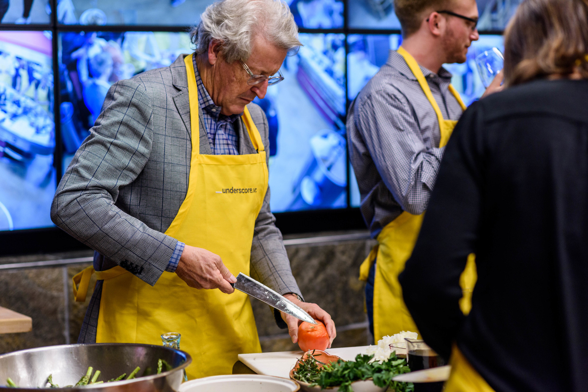 A man in a yellow apron chops a tomato while others participate in a cooking workshop.