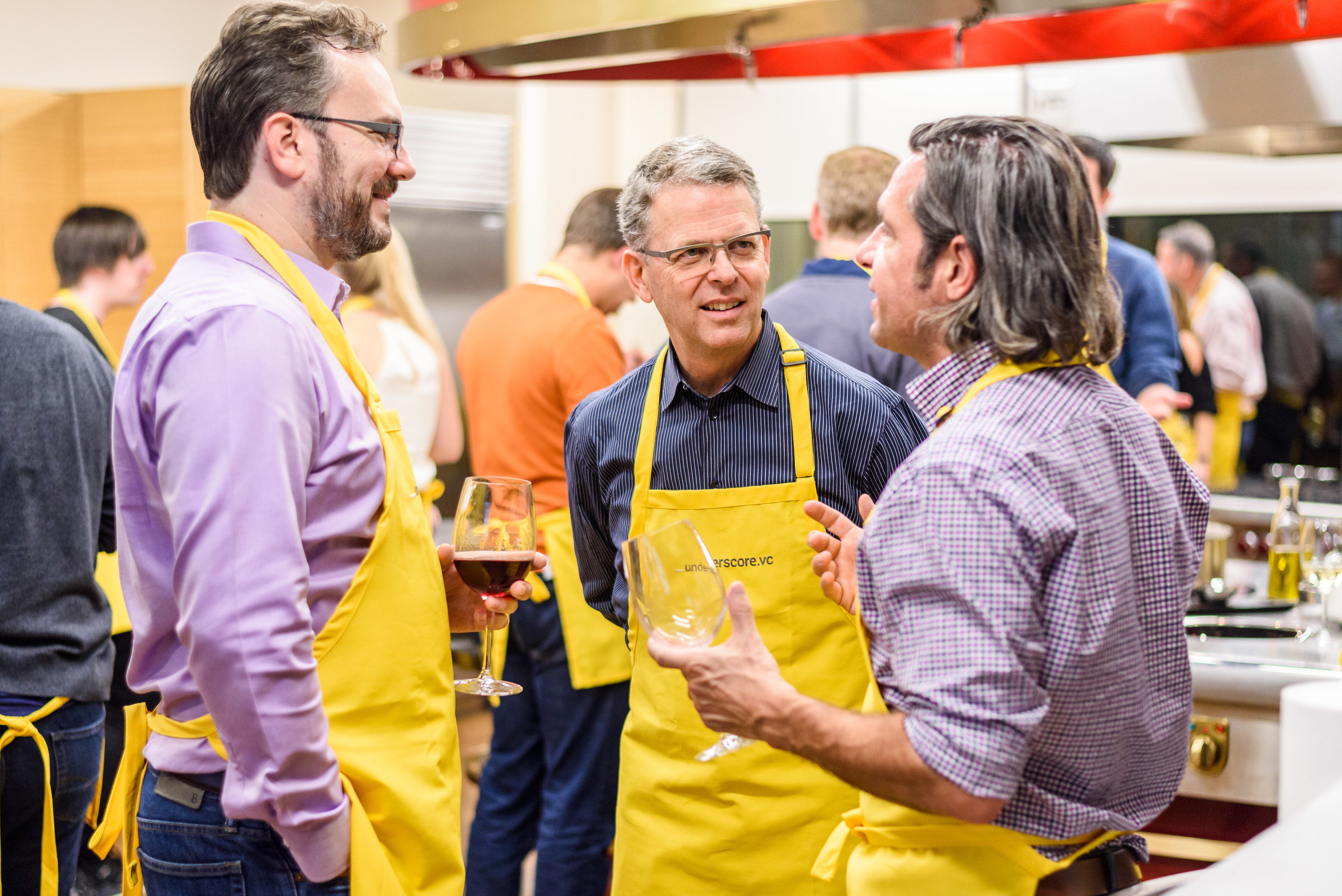 Three people wearing yellow aprons stand in a kitchen, holding drinks and talking during a cooking event.