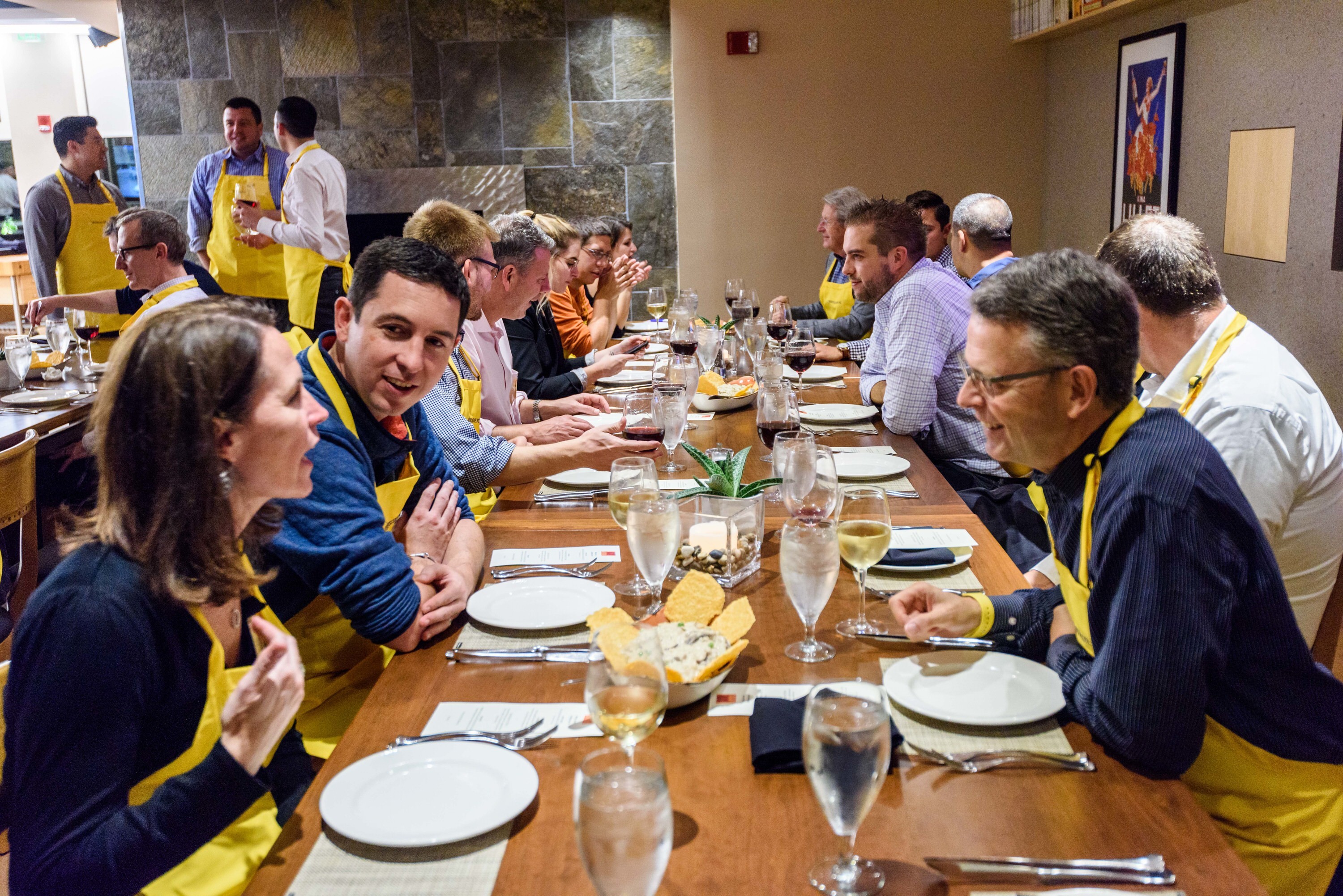A group of people wearing yellow aprons sit at a long table, talking and enjoying a meal together.