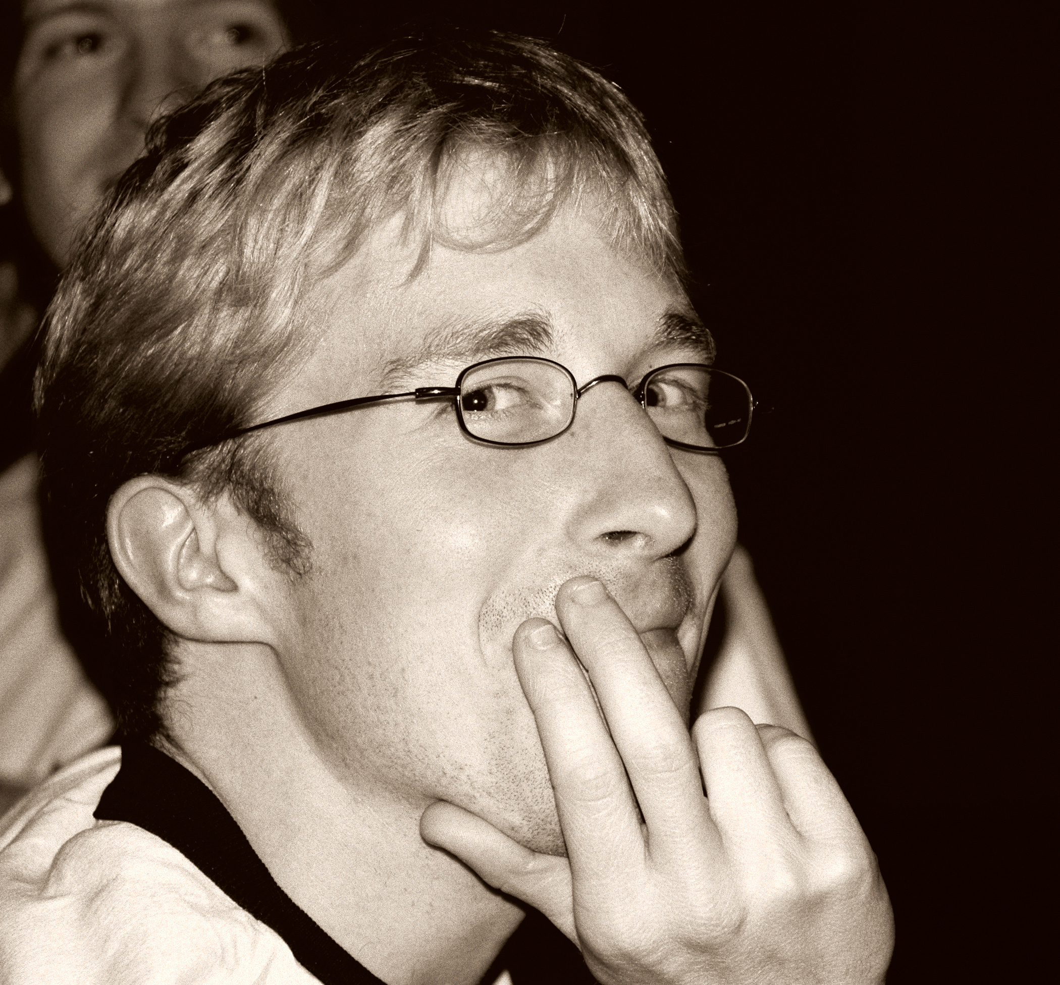 A person wearing glasses smiles playfully while resting their fingers on their lips at a social gathering.