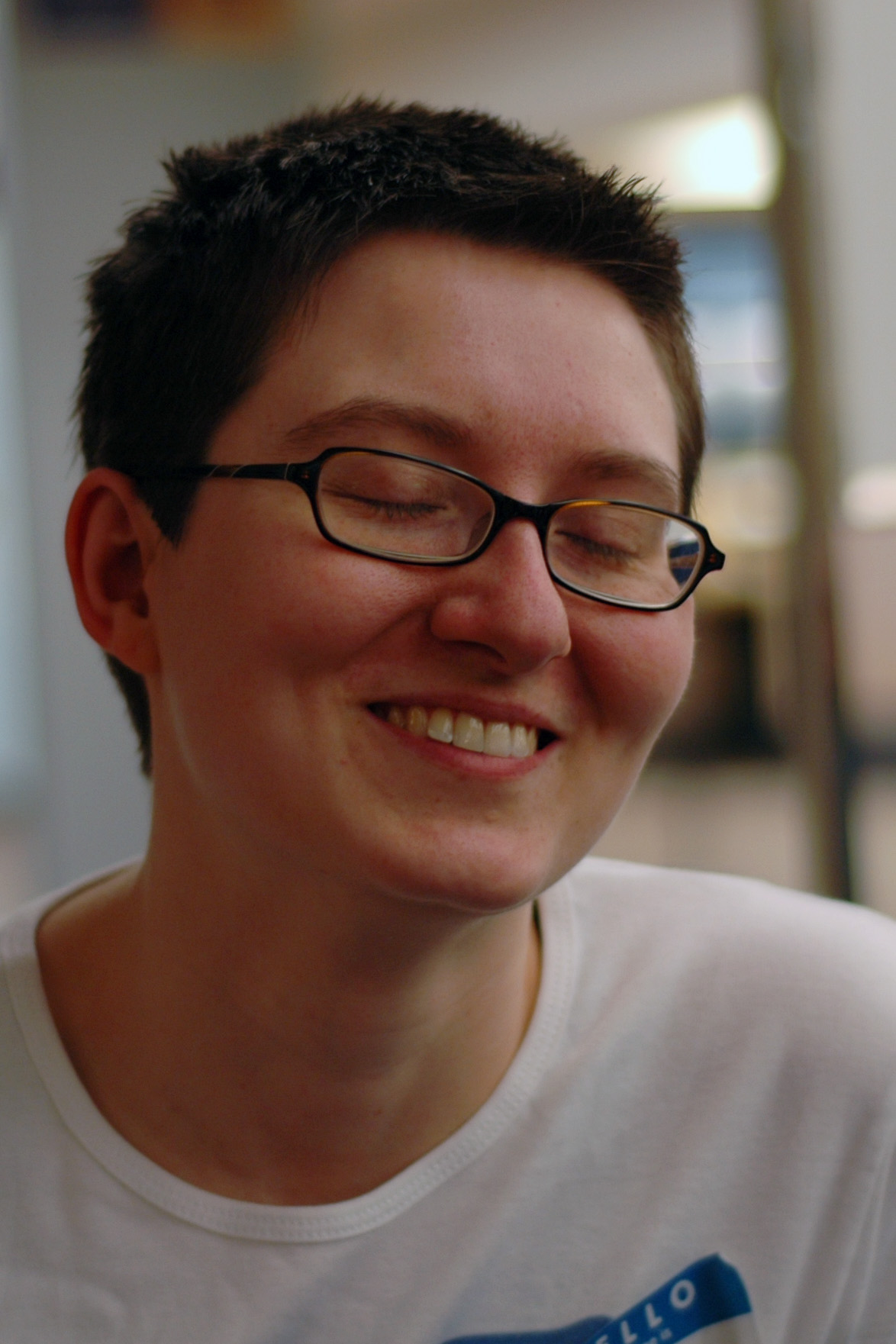 A person with short hair and glasses smiles with their eyes closed, wearing a white shirt with a name tag.