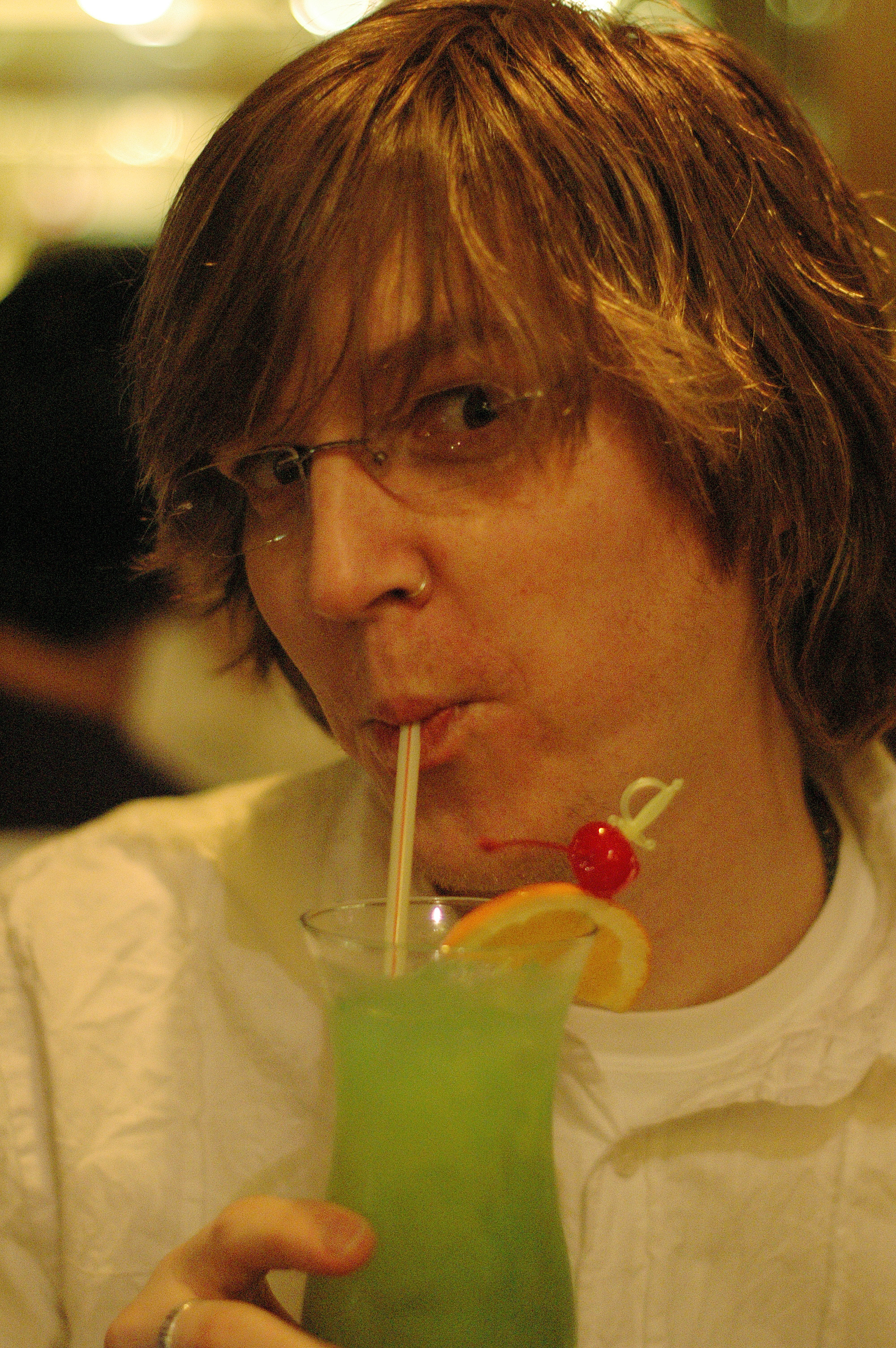 A person wearing glasses and a white shirt sips a green cocktail through a straw, garnished with fruit.