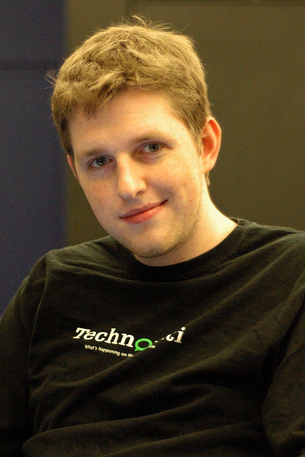 A man with short, light brown hair wearing a black Technorati T-shirt, smiling slightly while looking at the camera.