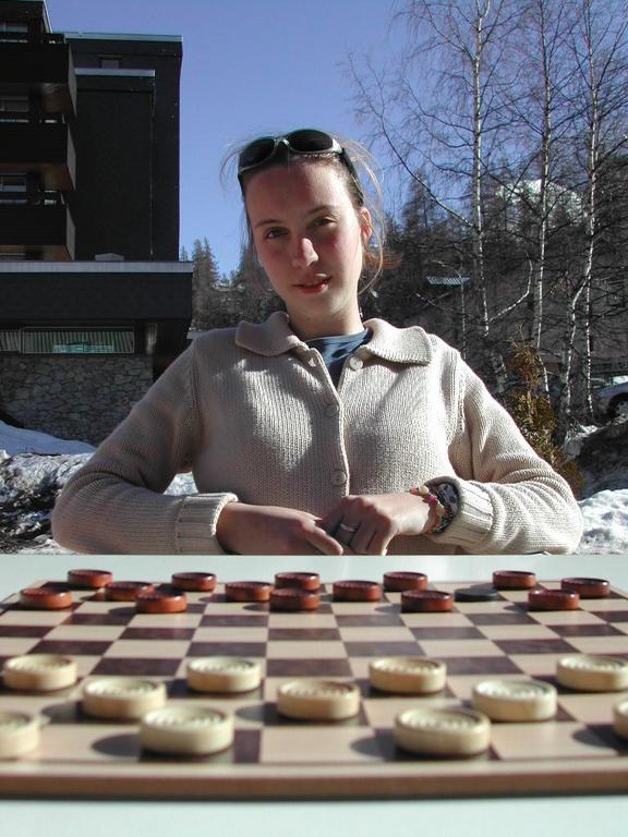 A person sits at an outdoor table, looking at a checkers board with game pieces arranged.