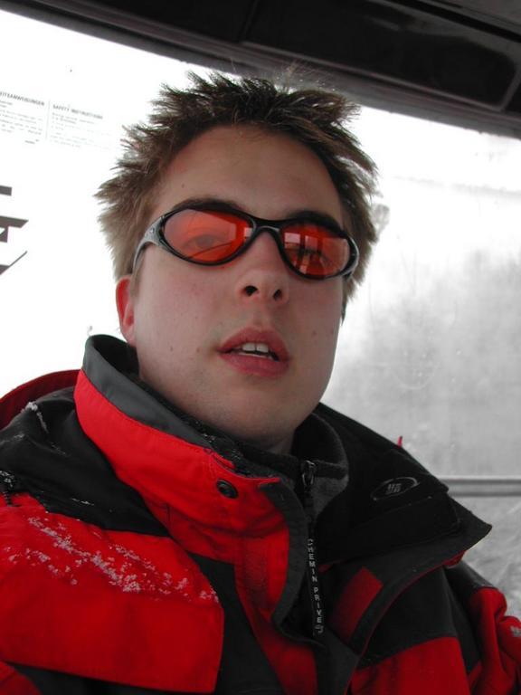 A person wearing red sunglasses and a red winter jacket takes a close-up selfie inside a ski lift.