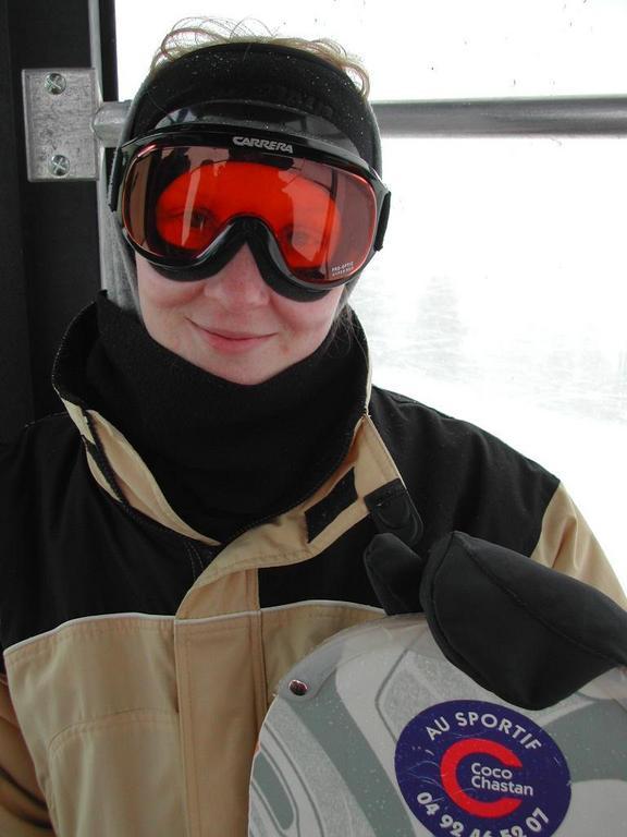 A person wearing ski goggles and winter gear holds a snowboard inside a ski lift or gondola.