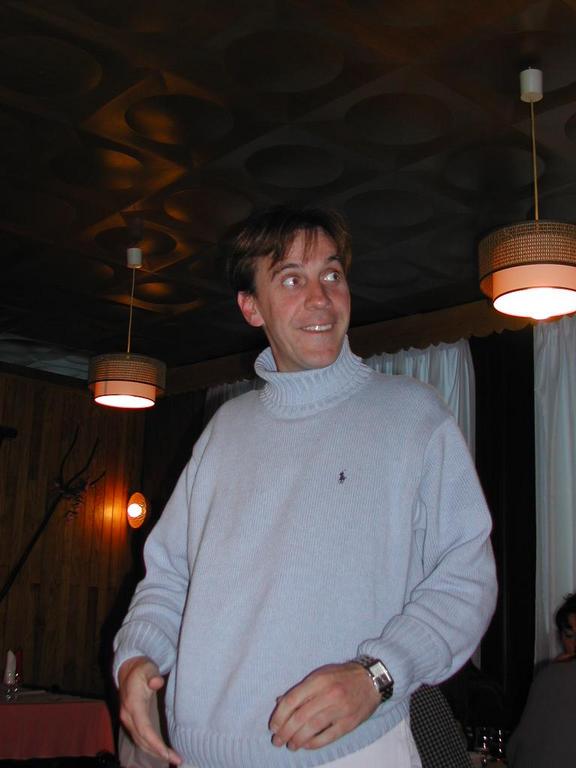 A man in a light-colored turtleneck sweater stands indoors, smiling and gesturing with his hands.