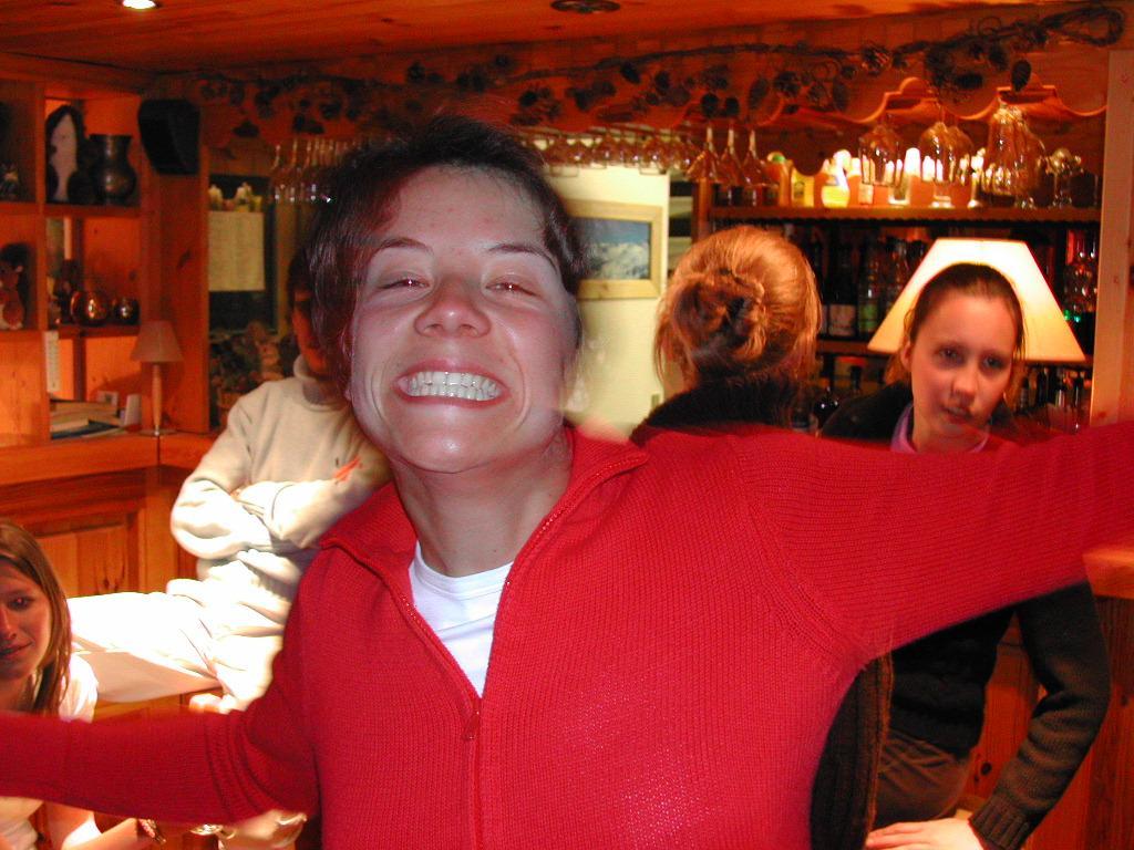 A person in a red sweater smiles widely with arms outstretched in a cozy bar setting.