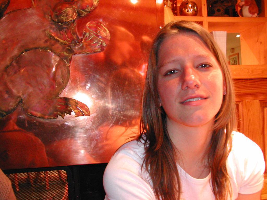A woman in a white shirt sits indoors, looking at the camera with a relaxed expression.