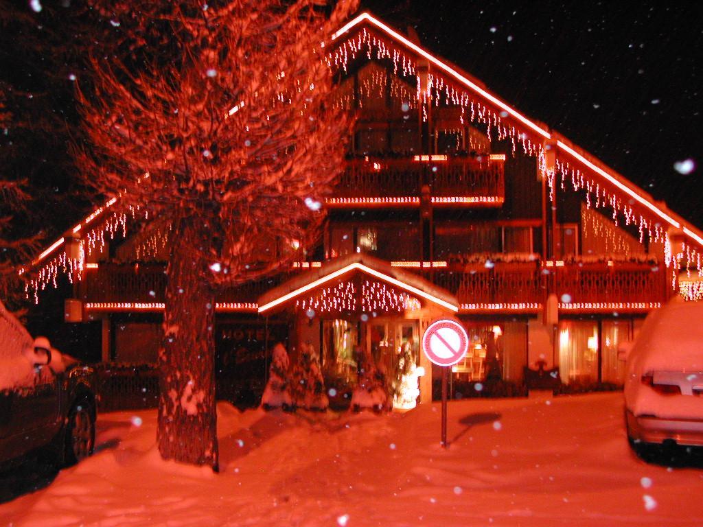 A chalet-style hotel decorated with red lights and icicle-shaped ornaments, surrounded by snow-covered trees and parked cars at night.