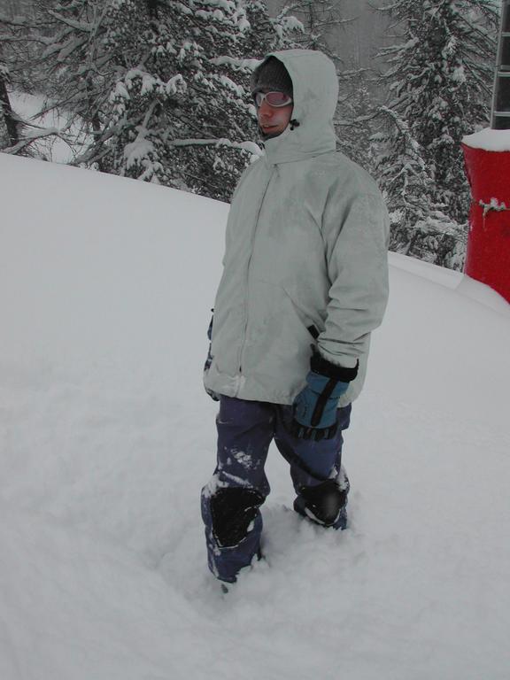A person in winter gear stands in deep snow, wearing a hooded jacket, gloves, and goggles.