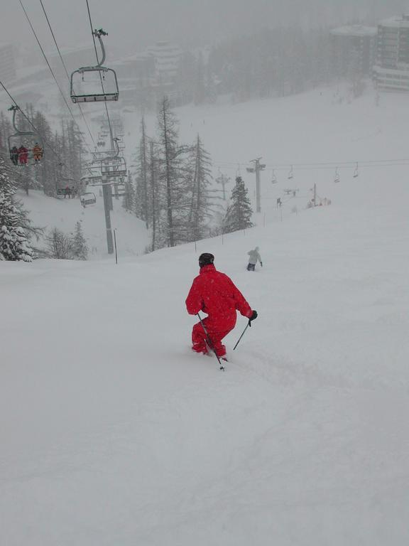 A skier in a red outfit moves downhill through deep snow, holding ski poles for balance.