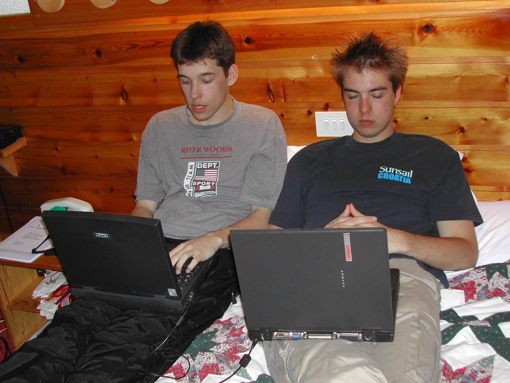 Two young men sitting on a bed using laptops, one typing while the other rests with hands clasped.