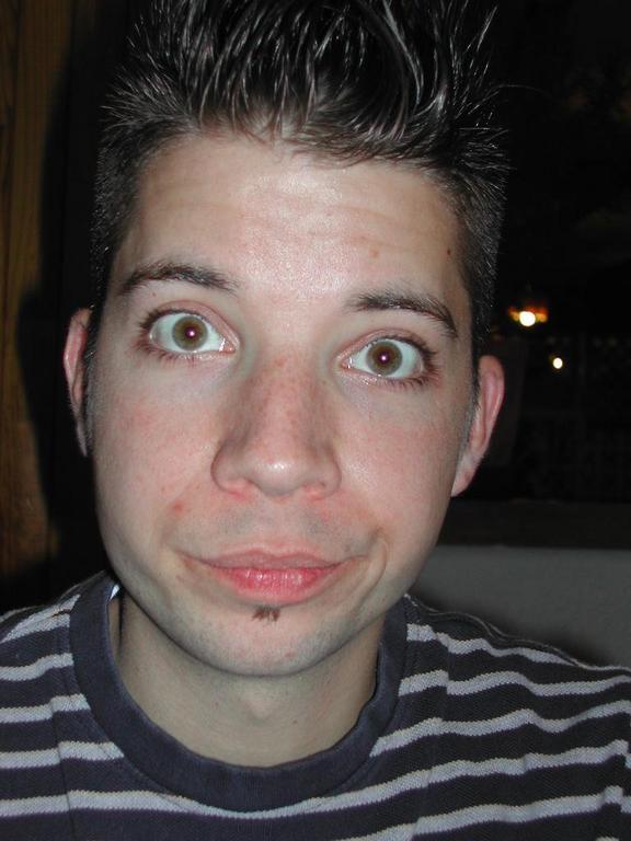 A person with spiky hair and a striped shirt makes a playful facial expression with wide eyes and a smirk.