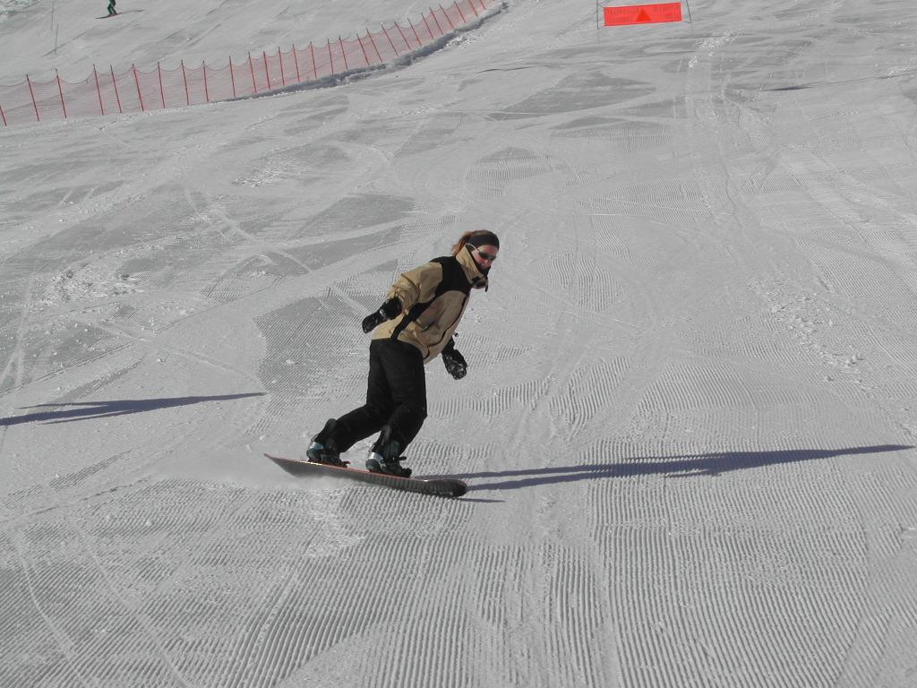 A person wearing a beige jacket and black pants snowboards down a groomed ski slope in sunny conditions.