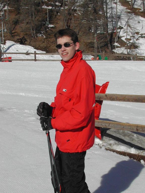 A person wearing a red jacket and sunglasses stands on a snowy slope, holding ski poles and smiling.