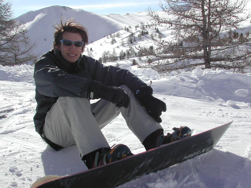 A person wearing winter gear sits on the snow with a snowboard, smiling at the camera.