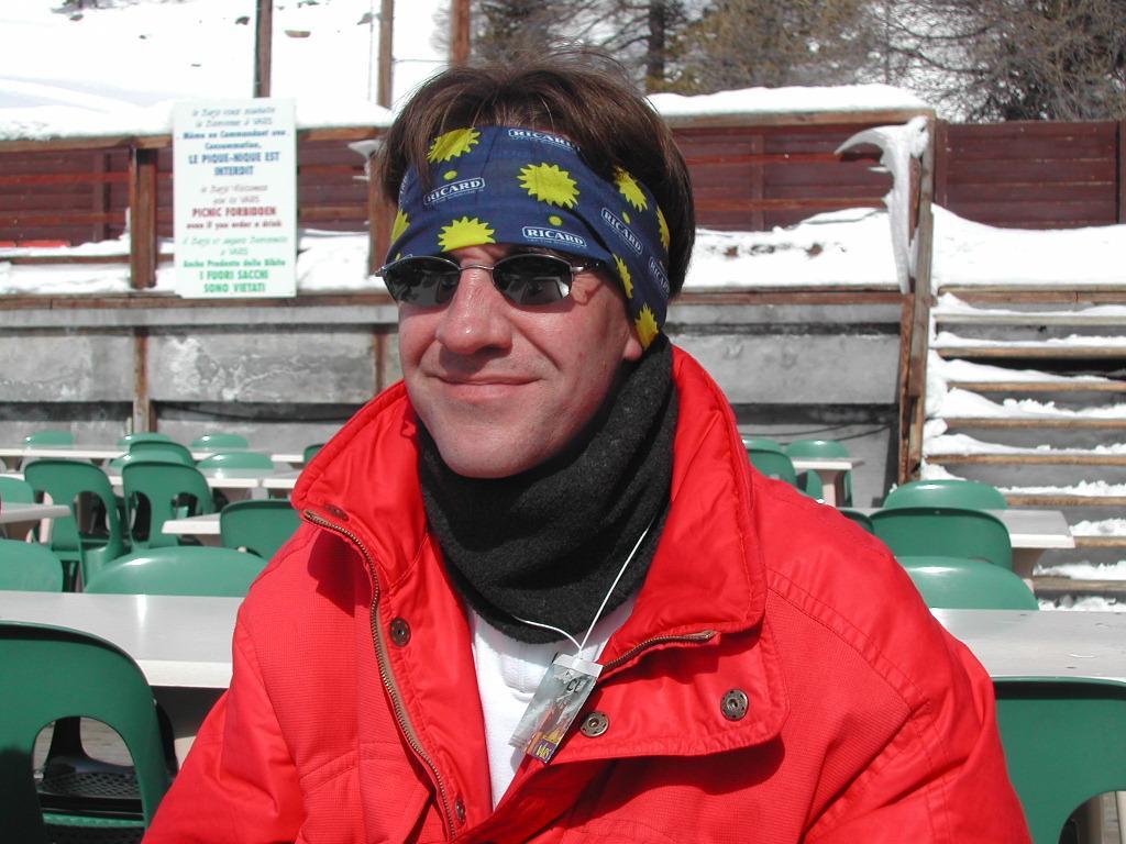 A person wearing sunglasses, a headband, and a red jacket sits outdoors in a snowy setting.