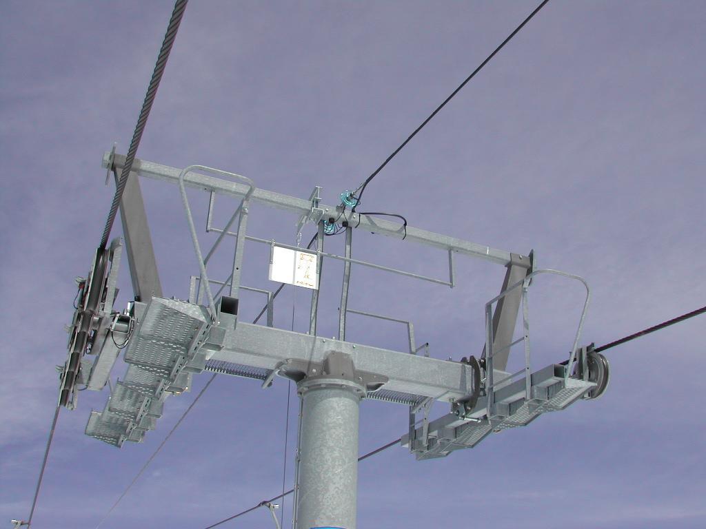 Close-up view of a ski lift support structure with cables and pulleys against a cloudy sky.