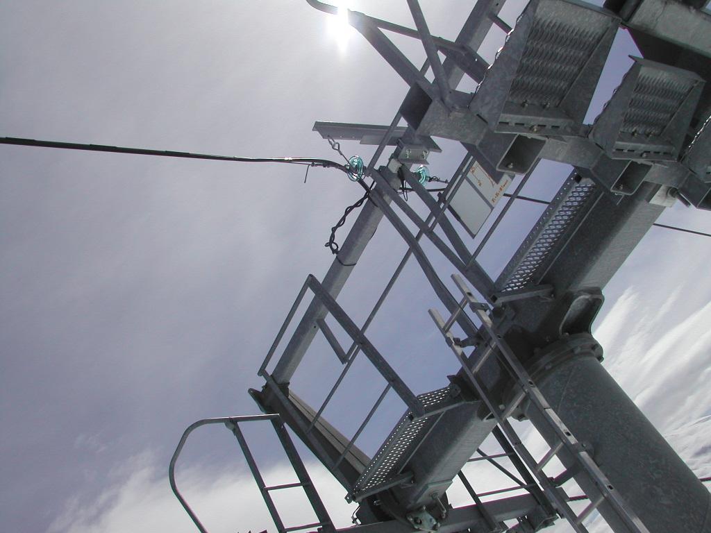 Metal ski lift structure with cables and pulleys, viewed from below against a cloudy sky.
