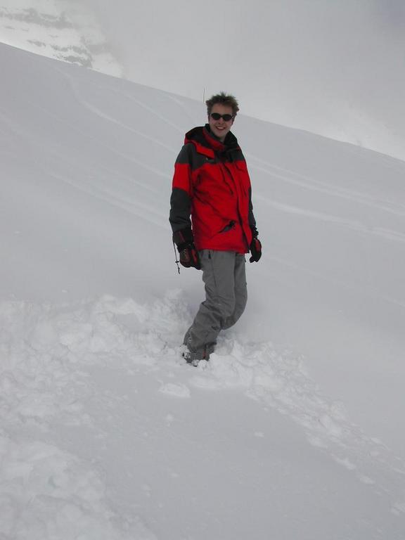 A person wearing a red jacket and sunglasses stands in deep snow on a snowy mountain slope.