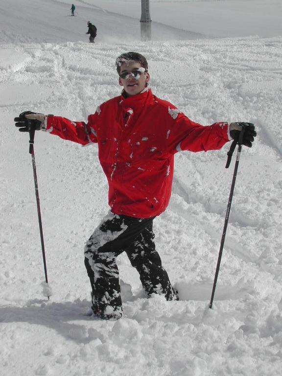 A person in a red jacket stands in deep snow, holding ski poles and covered in snow.