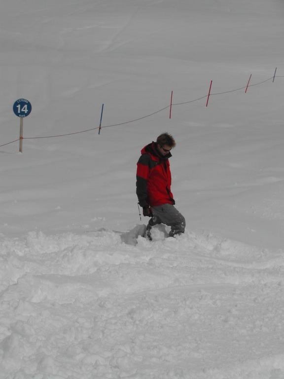 A person in a red jacket walks through deep snow on a ski slope, partially sinking with each step.