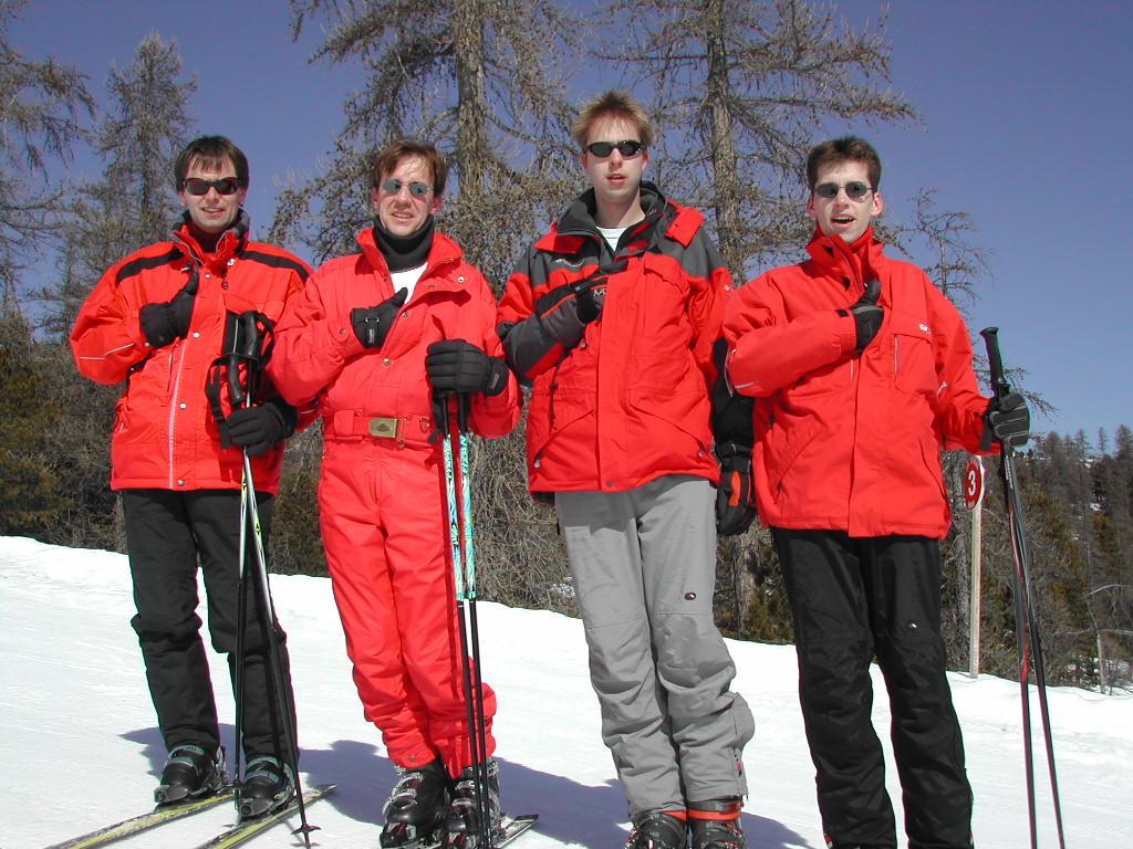 Four skiers in red jackets stand on a snowy slope, holding ski poles and wearing sunglasses.