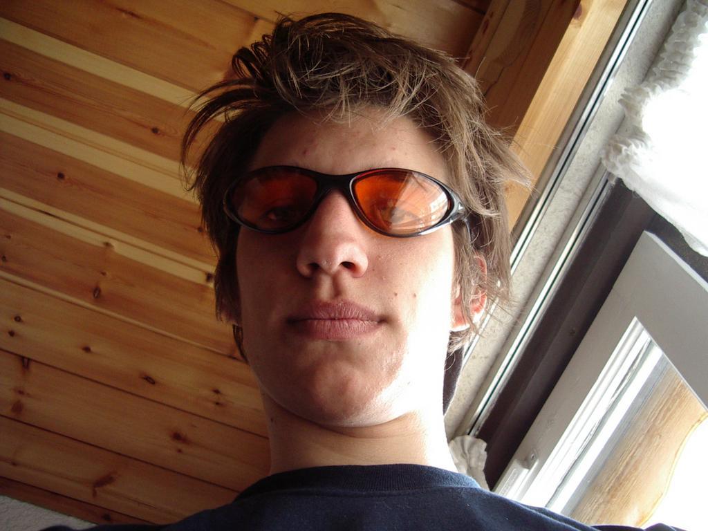 A person with messy hair and orange-tinted sunglasses looks down at the camera indoors near a window.