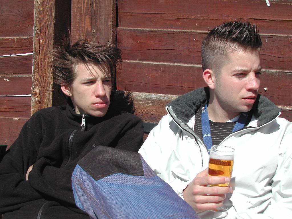 Two young men sit outside against a wooden wall.