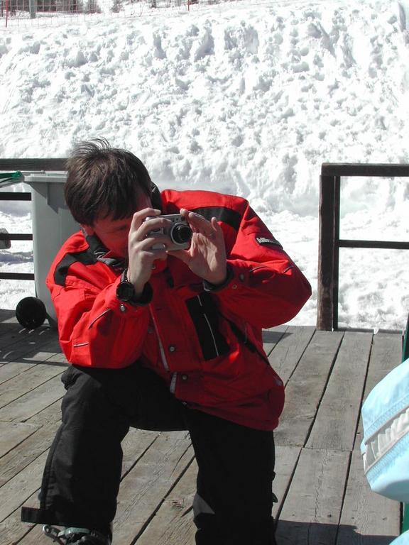 A person in a red jacket crouches and takes a photo with a small digital camera in a snowy setting.
