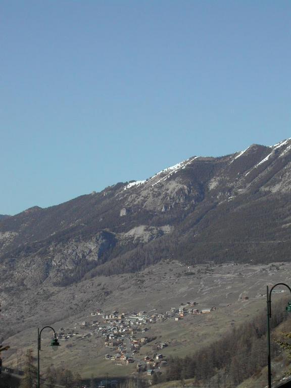 A small village is nestled on a hillside with towering mountains in the background under a clear blue sky.