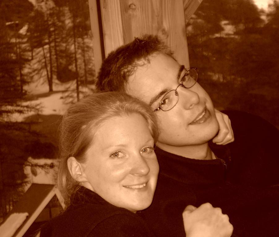 A smiling woman and a man wearing glasses sit closely together, embracing each other.