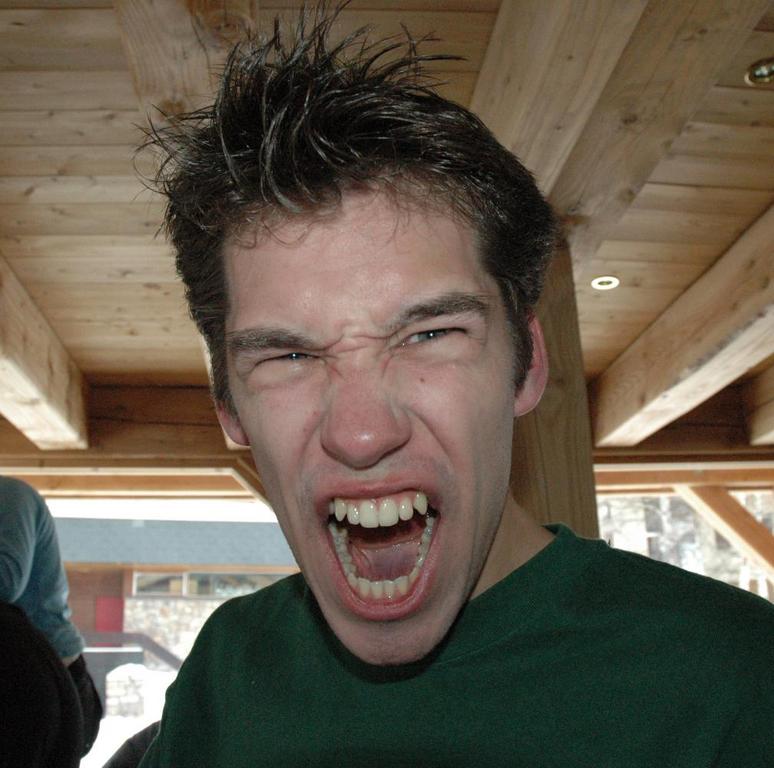 A young man with messy hair and a green shirt screams or makes a fierce expression indoors.