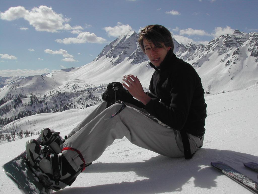 A person in snowboarding gear sits on a snowy slope, adjusting their gloves with mountains in the background.