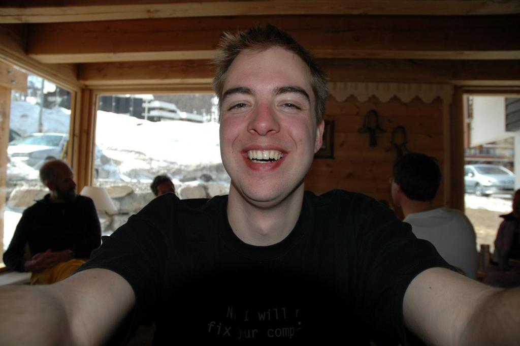 A person takes a close-up selfie while smiling in a wooden cabin with others sitting in the background.