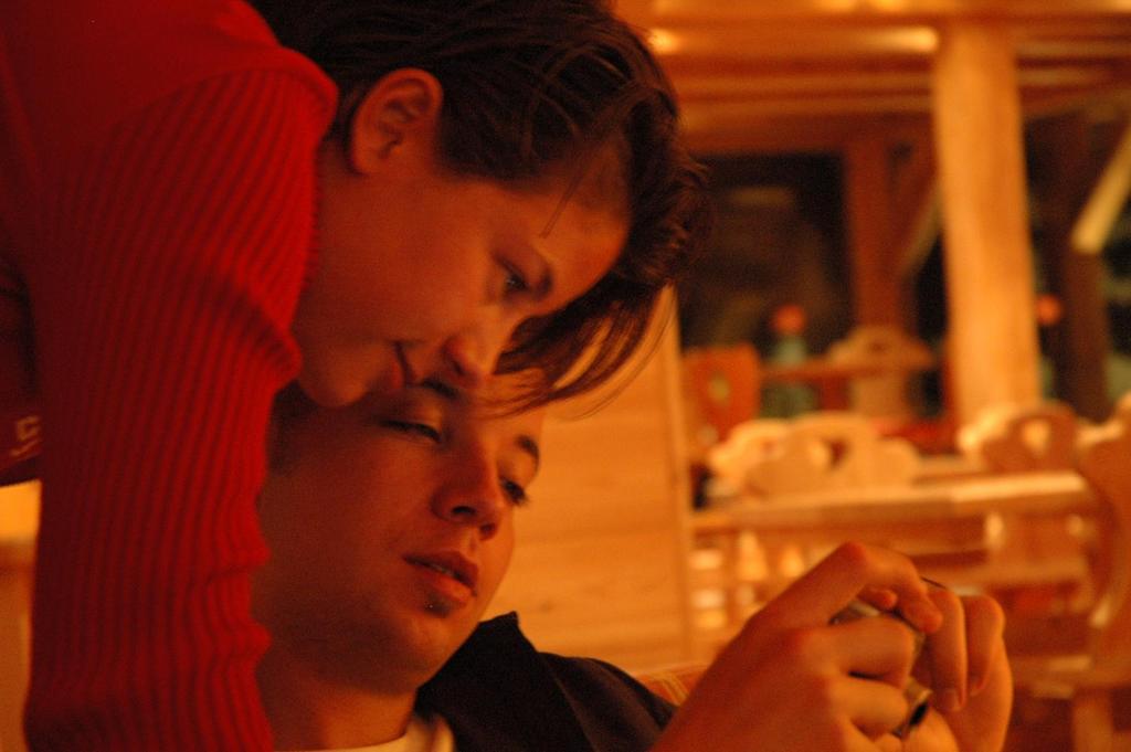 A woman leans over a man's shoulder as he focuses on a handheld device in a warm indoor setting.
