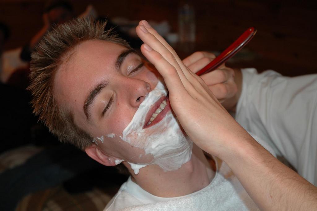 A person with shaving cream on their face is getting shaved with a straight razor while smiling.