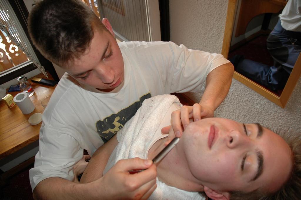 A person carefully shaves another person's face with a straight razor while they lie back with a towel around their neck.
