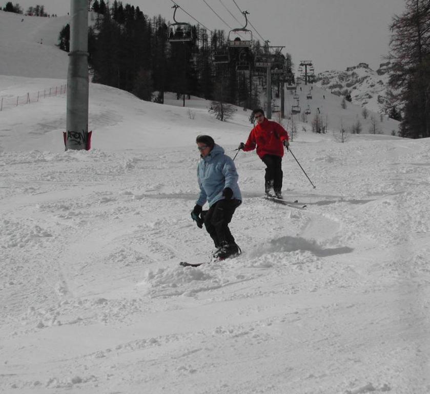 Two people skiing down a snowy slope, one in a blue jacket snowboarding and the other in a red jacket skiing.